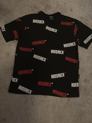 Svart HOODRICH t-shirt med tryck - Svart t-shirt från HOODRICH med korta ärmar och coolt all-over-tryck i vitt och rött. Oversized fit för en avslappnad streetwear-look. Tillverkad i mjuk bomull som känns skön mot huden. Perfekt för dig som gillar trendiga och bekväma plagg.