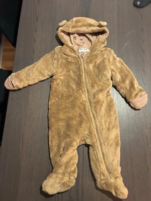 baby teddyoverall med öron - Mysig brun teddyoverall med huva och söta öron. Overallen har dragkedja framtill och mjukt, mönstrat innerfoder med ekollon och löv. Perfekt för kyliga dagar och supergosig att ha på bebis
