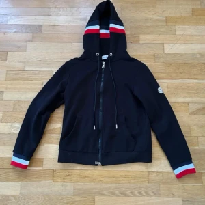 Svart hoodie från Moncler med dragkedja - Svart hoodie från Moncler med hel dragkedja, huva och snörning. Röd och vit rand på huva och ärmslut ger en sportig vibe. Moncler-logga på ärmen. Två fickor fram och ribbade muddar. Perfekt för dig som gillar stilrena detaljer.