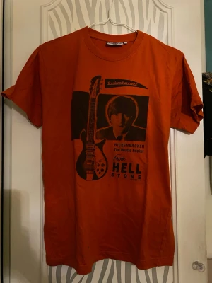 Orange Rickenbacker t-shirt med tryck - Cool orange t-shirt från Hellstone med stort svart tryck av en Rickenbacker-gitarr och siluett på framsidan. Beatles merch