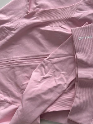 Rosa träningstights från DFYNE - Säljer ett par ljusrosa träningstights från DFYNE. Tightsen har hög midja, ribbade detaljer och är tillverkade i ett stretchigt material som passar perfekt för gymmet eller löpning. Snygg och bekväm passform med logga på benet.