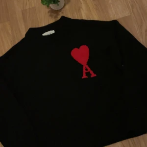 Svart stickad tröja från Ami Paris - Svart stickad tröja från Ami Paris med röd hjärt- och A-detalj på bröstet. Klassisk rund halsringning och ribbade muddar. Perfekt för dig som gillar stilrena plagg med en twist.