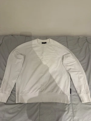 Vit sweatshirt från Emporio Armani - Vit sweatshirt från Emporio Armani med stort präglat logotypmönster på bröstet. Klassisk rund halsringning och ribbade muddar vid ärmslut och nederkant. Perfekt för en clean och stilren look.