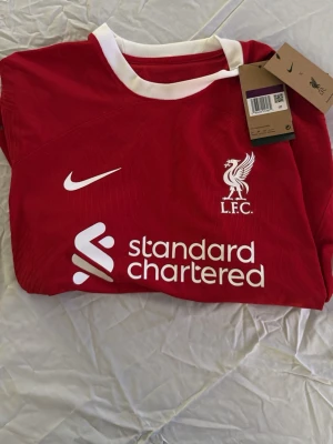 Liverpool matchtröja Nike röd - Säljer en röd Liverpool FC matchtröja från Nike med vita detaljer och klubbmärke på bröstet. Tröjan har tryck från Standard Chartered och Expedia samt vit krage och ärmslut. Perfekt för dig som älskar fotboll och vill stötta LFC.