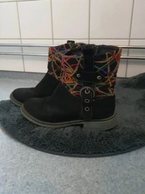 Svarta boots med färgglada detaljer - Unika svarta boots med färgglada broderade detaljer upptill och dekorativa spännen på sidan. Grov sula och dragkedja på insidan för enkel på- och avtagning. Perfekta för dig som vill sticka ut med din stil.