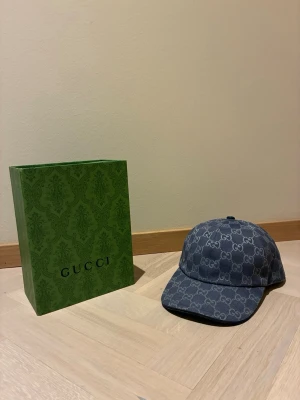 Blå denimkeps från Gucci - Säljer en blå keps från Gucci i denim med det klassiska GG-monogrammet över hela kepsen. Kepsen har böjd skärm och justerbar rem bak. Insidan är fodrad i ljus bomull och etiketten visar äkthet. Boxen från Gucci medföljer.