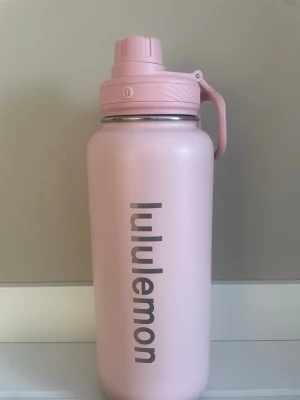 Rosa vattenflaska från Lululemon - Säljer en stilren rosa vattenflaska från Lululemon med praktiskt handtag och skruvlock. Flaskan är tillverkad i rostfritt stål och har Lululemon-loggan tryckt i silver på sidan. Perfekt för träning eller vardagsbruk. Svart tygpåse med Lululemon-logga medföljer. Den är 30cm hög och rymmer mycket dryck 🩷