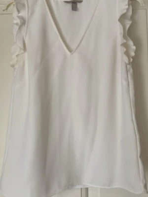 Vit volangblus från H&M - Säljer en vit blus från H&M med v-ringning och volangdetaljer längs axlarna. Blusen är ärmlös och har en luftig passform, perfekt för varma dagar. Materialet känns mjukt och lätt mot huden.