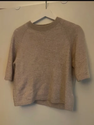 Beige stickad kortärmad tröja - Mysig beige stickad tröja med korta ärmar och rund halsringning. Tröjan har en avslappnad passform och ribbad kant vid halsen. Perfekt för lager på lager och enkel att matcha med olika stilar.