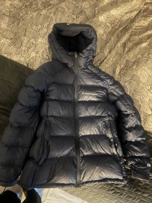 Mörkblå dunjacka Peak Performance - Snygg mörkblå dunjacka från Peak Performance med huva och dragkedja framtill. Jackan har två sidofickor och en bröstficka med logga. Puffig modell som håller dig varm under kalla dagar. Perfekt för vinter och outdoor.
