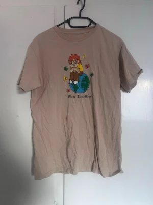Beige t-shirt Bless This Mess - Beige t-shirt från Butter Goods med tryck av en tecknad figur som sitter på en jordglob, omgiven av fjärilar och texten 'Bless This Mess'. T-shirten har rund hals och korta ärmar. Perfekt för dig som gillar streetwear och coola prints.