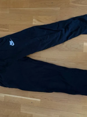Svarta Nike mjukisbyxor - Säljer ett par svarta mjukisbyxor från Nike med klassisk vit logga på vänster lår. Byxorna har en avslappnad passform och är tillverkade i mjukt material, perfekta för chill dagar. Resår i midjan och vid benslut för extra komfort.