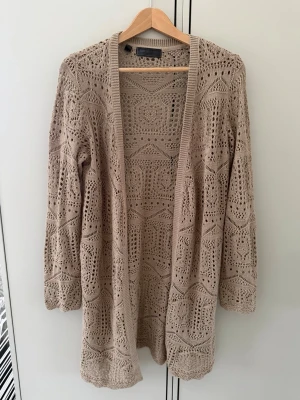 Beige virkad kofta med mönster - Lång, öppen beige kofta med virkat mönster och trekvartsärmar. Koftan har en avslappnad passform och är tillverkad i ett lätt, luftigt material som passar perfekt för lager på lager. Snygg detaljrik design som ger en bohemisk känsla.