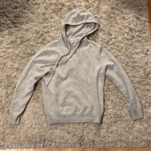 Kashmir hoodie - Säljer en grå kashmir hoodie från St studios med klassisk huva och dragsnören. Köpt för 1500kr säljer för 950kr.Hoodien har långa ärmar och ribbade muddar vid ärmslut och nederkant. Tillverkad i mjukt material som känns skönt mot huden. Perfekt för en avslappnad och trendig look. Har gått lite sönder vid kragen som syns på bilden.