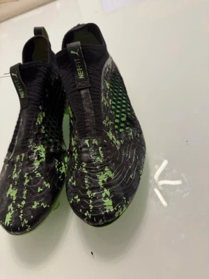 Puma Future Z svarta/gröna fotbollsskor - Säljer ett par Puma Future Z fotbollsskor i svart med gröna detaljer och mönster. Skorna har en strumpliknande passform utan snörning, meshpaneler på sidorna och greppvänlig yttersula. Perfekta för dig som vill sticka ut på planen! Skorna känns som storlek 43