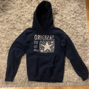 Original hoodie - Mörkblå hoodie från Original Fit med stor huva och magficka. Tryck på bröstet med texten 'ORIGINAL FIT EST. 2024' och en stor vit stjärna omgiven av mindre stjärnor. Mjuk och skön, perfekt för chill dagar. Klassisk passform och ribbade muddar.