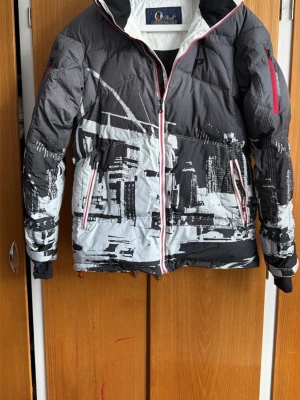 Svart och vit pufferjacka med stadsmotiv - Säljer en svart och vit pufferjacka från Peak Mountain med coolt stadsmotiv och röda dragkedjor. Jackan har huva, två framfickor och en ficka på ärmen. Perfekt för kalla vinterdagar och riktigt snygg streetstil.