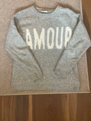 Grå stickad tröja med text AMOUR - Mysig grå stickad tröja med vit text 'AMOUR' framtill. Tröjan har rund halsringning, långa ärmar och ribbstickade muddar vid ärmslut och nederkant. Perfekt för dig som gillar en avslappnad och trendig stil.