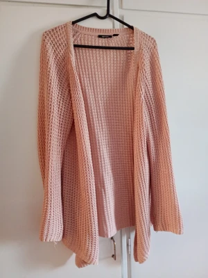 Ljusrosa stickad öppen kofta - Säljer en ljusrosa stickad kofta med öppen front och lång ärm. Koftan har ett luftigt, grovmaskigt mönster och ribbade muddar. Perfekt att slänga över en topp för en avslappnad look.