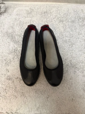 Svarta ballerinaskor i mesh och skinn - Säljer ett par svarta ballerinaskor med ovandel i både mesh och skinnimitation. Skorna har en enkel, rundad tå och diskreta röda detaljer på insidan. Perfekta för dig som gillar en sportig men stilren look.
