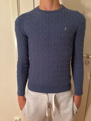 Blå kabelstickad tröja från Morris  - Snygg blå kabelstickad tröja från Gant med rund halsringning och diskret logga broderad på bröstet. Tröjan har långa ärmar och ribbade muddar vid ärmslut och nederkant. Perfekt för en stilren och klassisk look. STORLEK S MEN DEN EÄR KRYMPT PASSAR MER SOM XS. Killen på bilden är 172 cm och väger 55kg så passar dig runt 50kg och 160-165 cm lång