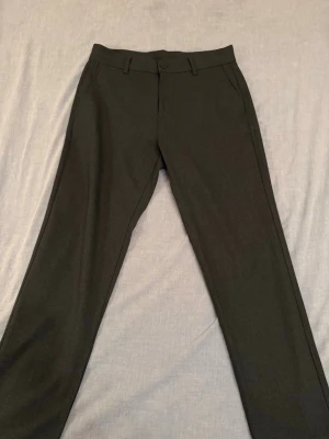 Chinos Bekväm Struktur - Snygga mörkgrå chinos med rak passform och diskreta sidfickor. Byxorna har hällor för bälte och knäppning fram. Perfekta för en stilren look. Materialet känns mjukt och följsamt, en blandning av polyester, viskos och elastan. Endast använd 2 gånger. Storlek 40