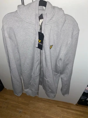 Ljusgrå zip hoodie från Lyle & Scott - Ljusgrå hoodie med dragkedja från Lyle & Scott. Klassisk design med huva och fickor framtill. Märkets ikoniska gula logga broderad på bröstet. Tillverkad i mjukt material som passar perfekt till en avslappnad stil.