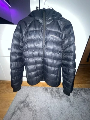 Svart pufferjacka Peak Performance - Svart pufferjacka från Peak Performance med huva och dragkedja framtill. Jackan har quiltad design med breda kanaler och diskret logga på bröstet. Perfekt för kalla dagar och enkel att matcha med olika outfits.