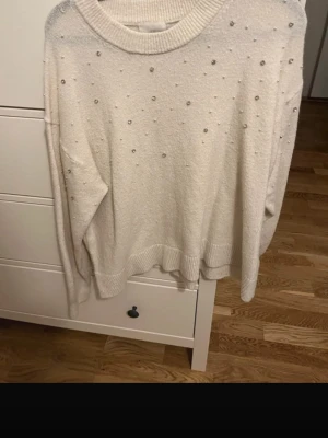 Vit stickad tröja med pärlor - Säljer en vit stickad tröja med rund hals och dekorativa små pärlor över hela framsidan. Tröjan har långa ärmar och ribbade muddar vid ärmslut och nederkant. Lite nopprig vid armhålor men syns ej tydligt