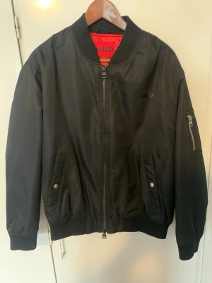 Svart bomberjacka från HUGO - Snygg svart bomberjacka från HUGO med klassisk ribbad krage, muddar och nederkant. Jackan har dragkedja framtill, två snedställda fickor med tryckknapp och en ärmficka med dragkedja. Röd innerfoder och diskret HUGO-logga på bröstet.