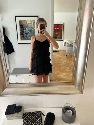 Svart volangklänning med smala axelband - Säljer en svart kort klänning med smala axelband och flera volangdetaljer. Klänningen har en lätt och luftig känsla, perfekt för varma dagar. Enkel och stilren design som ger en trendig look.