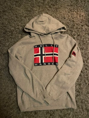 Grå Helly Hansen hoodie med flagga - Snygg grå hoodie från Helly Hansen med stor norsk flagga och logga på bröstet samt HH-märke på ärmen. Klassisk huva med dragsko och känguruficka framtill. Tillverkad i mjuk bomullsblandning, perfekt för chill dagar.