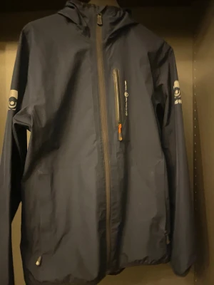 Mörkblå vindjacka Sail Racing - Snygg mörkblå vindjacka från Sail Racing med huva och hel dragkedja framtill. Jackan har en bröstficka med vattentät dragkedja och två sidofickor. Logotyp på ärmen och bröstet. Tillverkad i lätt och tåligt material, perfekt för blåsiga dagar. Ny pris ligger på ungefär 2800