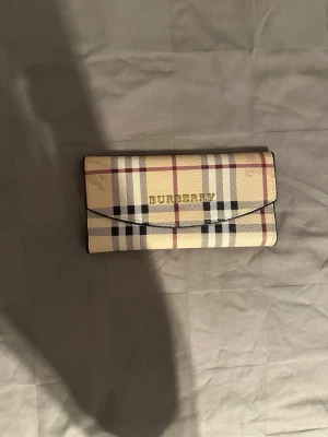 Burberry plånbok i klassiskt rutmönster - Säljer en snygg plånbok från Burberry med det ikoniska beige, svarta och röda rutmönstret. Plånboken har guldfärgad Burberry-logga på framsidan och är tillverkad i tåligt material med slät yta. Perfekt för dig som gillar klassisk och tidlös design.