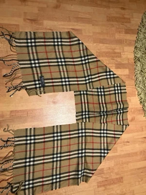 Beige rutig halsduk från Burberry - Klassisk beige halsduk från Burberry med det ikoniska rutmönstret i svart, vitt och rött. Halsduken har fransar i ändarna och är tillverkad i mjuk ull. Perfekt accessoar för att lyfta din outfit med en tidlös och lyxig känsla.