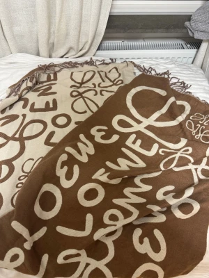 Brun och beige halsduk från Loewe - Säljer en stor, mjuk halsduk från Loewe i brunt och beige med ikoniskt logomönster och fransar i kanterna. Perfekt accessoar för att lyfta din outfit med en lyxig touch och trendig design.