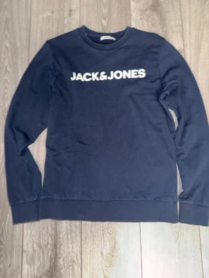 Mörkblå sweatshirt Jack & Jones - Mörkblå sweatshirt från Jack & Jones med vit logga tryckt över bröstet. Klassisk rund halsringning och ribbade muddar vid ärmslut och nederkant. Perfekt för en avslappnad och stilren look.