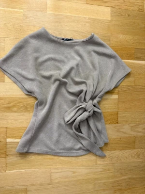 Beige topp med knyt från Zara - Snygg beige topp från Zara med korta ärmar och rund halsringning. Toppen har en unik knyt-detalj i sidan som ger en cool och trendig look. Perfekt att styla till jeans eller kjol för en avslappnad men chic outfit.