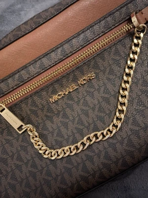 Michael Kors brun handväska med kedja - Snygg handväska från Michael Kors i brunt konstläder med logomönster och detaljer i ljusbrunt. Väskan har guldfärgad kedja och dragkedja framtill samt Michael Kors-logga i guld. Perfekt accessoar för att lyfta din outfit.