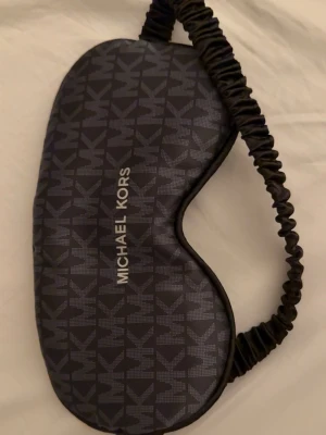 Svart sovmask från Michael Kors - Snygg svart sovmask från Michael Kors med elastiskt band och MK-logga över hela framsidan. Masken är mjuk och bekväm, perfekt för att blockera ljus när du vill vila. Utsidan har ett stilrent mönster och insidan är slät och skön mot huden.