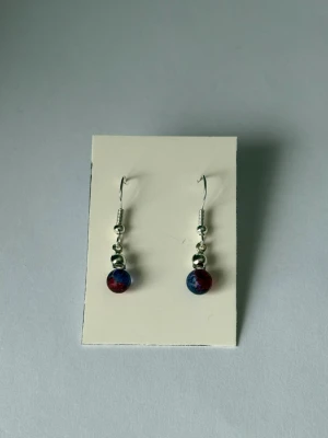Blå-röda pärlörhängen - Söta örhängen med silverfärgade krokar och detaljer.                                                                 Pärlan är blå-röd. 💫                                Örhängerna är självklart helt oanvända och de är nickelfria.                                                                    De sitter på en bit kartong för att enklare kunna visa hur de ser ut.                                                     Ha det fint!🌻