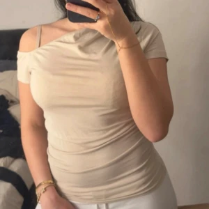 Beige oneshoulder topp i bomull - Trendig beige topp med oneshoulder-design och rynkade sidor. T-shirten är tight och har kort ärm på ena sidan, perfekt för en cool och avslappnad look. Tillverkad i mjuk bomull som känns skön mot huden.