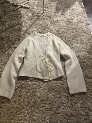 Krämvit omlottkofta med knytband - Säljer en krämvit omlottkofta med långa ärmar och knytband i midjan. Koftan har en mjuk och lättstickad känsla, perfekt för lager på lager. Enkel och stilren design som passar till det mesta.