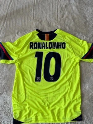  FC Barcelona Ronaldinho tröja - Säljer en gul FC Barcelona matchtröja från Nike med Ronaldinho och nummer 10 på ryggen (2005-version). Tröjan har klubbmärke på bröstet, svarta detaljer och färgade ränder på ärmarna. Perfekt för dig som älskar fotboll och vill ha en fin tröja till vår/sommar. Oanvänd. 