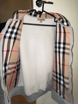 Grå hoodie från Burberry med dragkedja - Säljer en grå hoodie från Burberry med klassiskt rutigt foder i huvan och insidan. Hoodien har dragkedja framtill, fickor och en broderad Burberry-logga på bröstet. Materialet är mjukt och bekvämt, perfekt för en avslappnad stil.