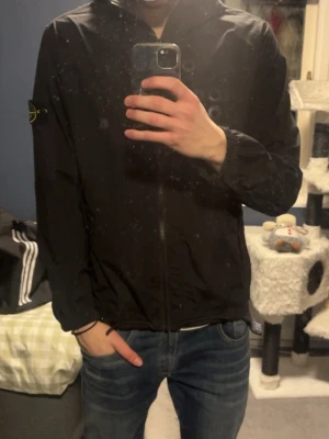 Svart vindjacka från Stone Island - Säljer en svart vindjacka från Stone Island med klassisk logotyp-patch på vänster ärm. Jackan har dragkedja framtill, huva och elastiska muddar vid ärmslut och midja. Perfekt för dig som vill ha en stilren och sportig look.