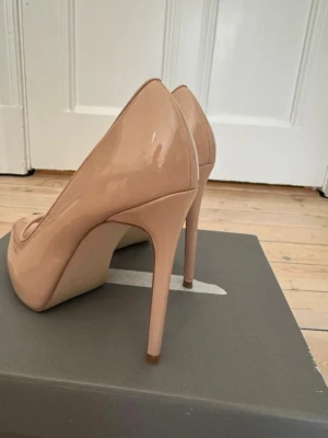 Nude peep toe pumps från Zara - Snygga nude-färgade pumps från Zara med peep toe och hög klack. Skorna är i glansigt lackmaterial som ger en elegant look. Perfekta för dig som vill ha ett par klassiska och stilrena klackar till garderoben.