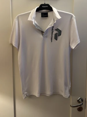 Vit pikétröja från Peak Performance - Klassisk vit pikétröja från Peak Performance med korta ärmar och krage. Tröjan har en diskret logga och märke på bröstet samt knappslå vid halsen. Tillverkad i mjuk bomull som känns skön mot huden. Perfekt för en sportig och stilren look.