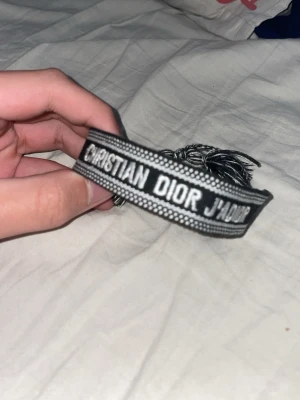Christian Dior J'adior tygarmband - Snyggt svart och vitt tygarmband från Christian Dior med texten 'J'adior' broderad. Armbandet har dekorativa tofsar i ändarna och justerbar knytning. Perfekt accessoar för att lyfta din outfit med en exklusiv touch.