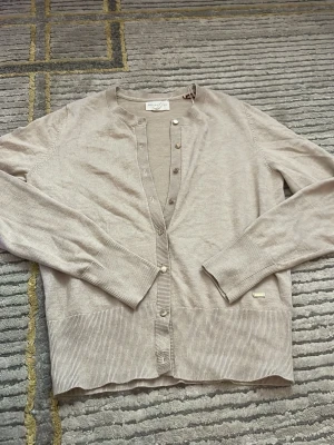 Beige kofta från Holly & Whyte by Lindex  - Stilren beige kofta från Holly & Whyte by Lindex med rund halsringning och knappar framtill. Tröjan har långa ärmar och ribbade muddar samt nederkant. Perfekt att slänga över en t-shirt eller linne för en avslappnad look.